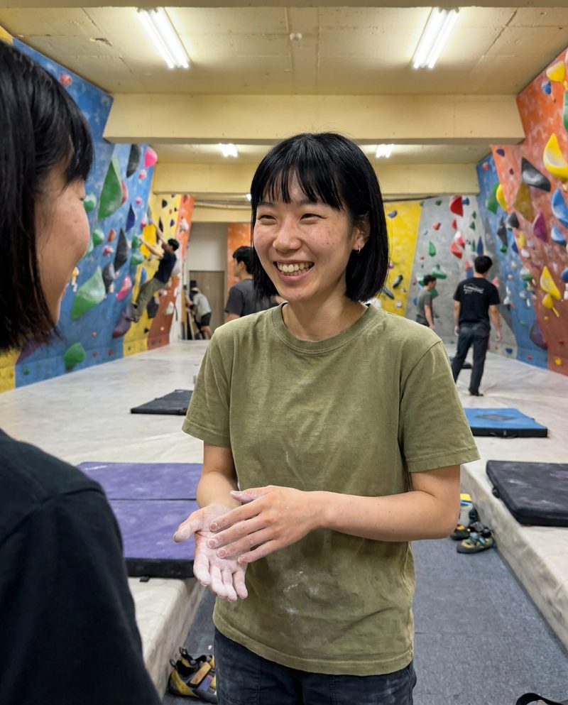 Chalking her hands at a bouldering gym in the evening, overhead white lights, grinning at a friend, waist-up.