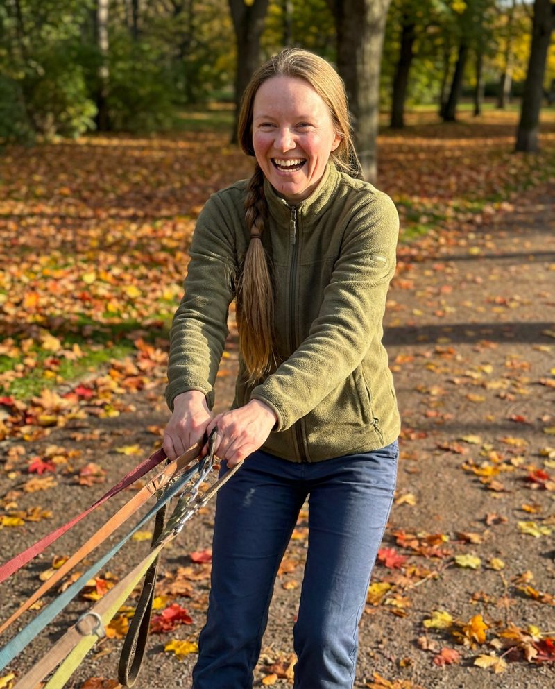 Holding five leashes on a park path in the morning, autumn leaves, low sun from the right, being pulled forward and laughing, full figure.