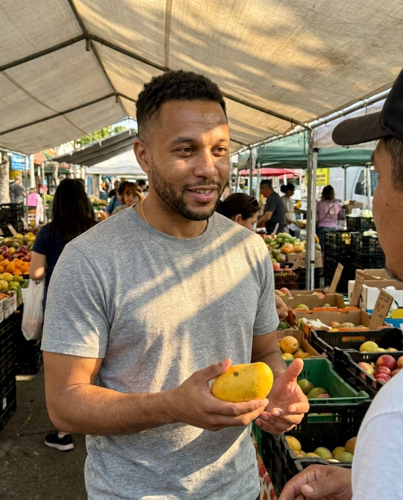 Buying fruit at a busy open-air market in the morning, dappled light through a canvas awning, holding a mango and talking with the vendor, waist-up.