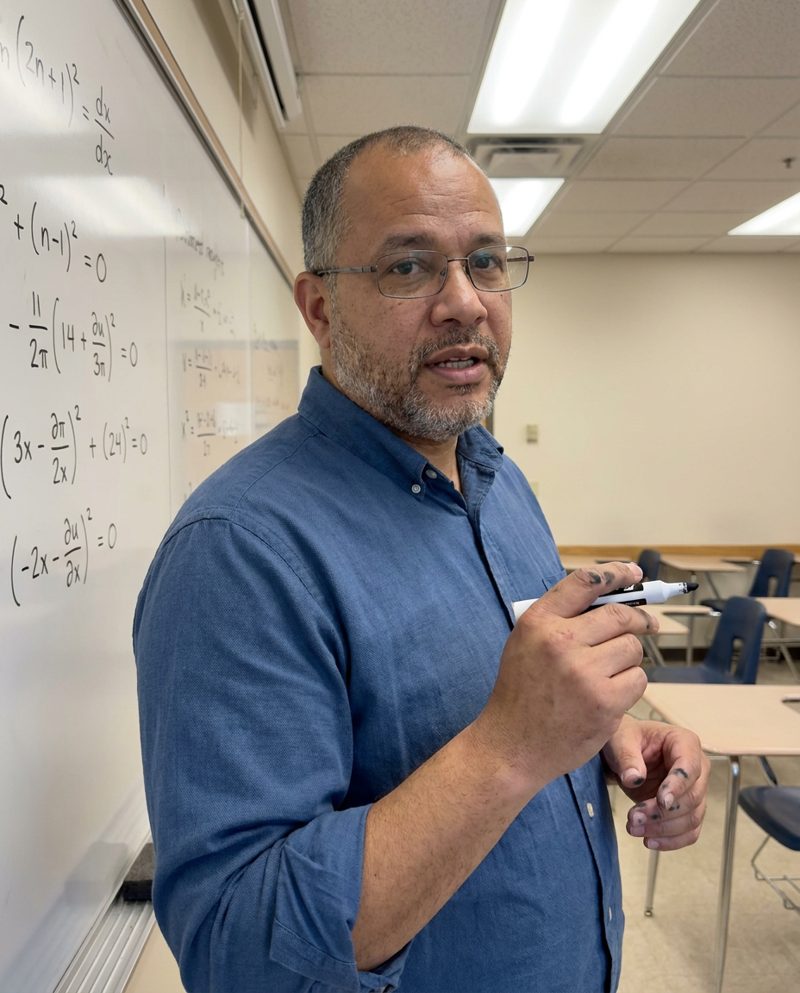 Writing an equation on a whiteboard in a classroom, overhead fluorescent light, turning to the class mid-sentence with a marker in hand, waist-up.