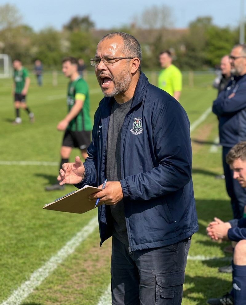 Holding a clipboard on the sideline of a football pitch on a bright Saturday morning, sun from the left, shouting encouragement, three-quarter body.